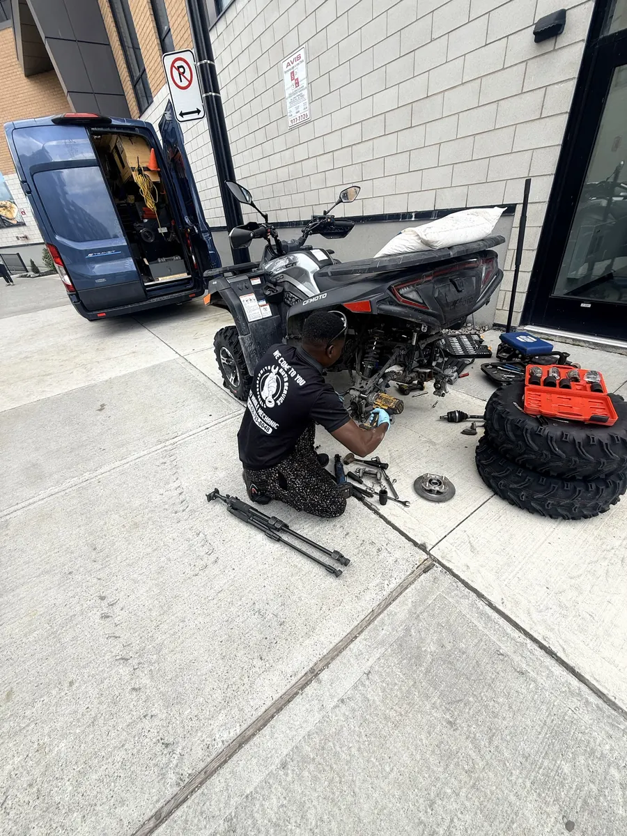 Elie repairing the rear hub of a CFMoto ATV on the sidewalk, blue Ford Transit service van open behind him