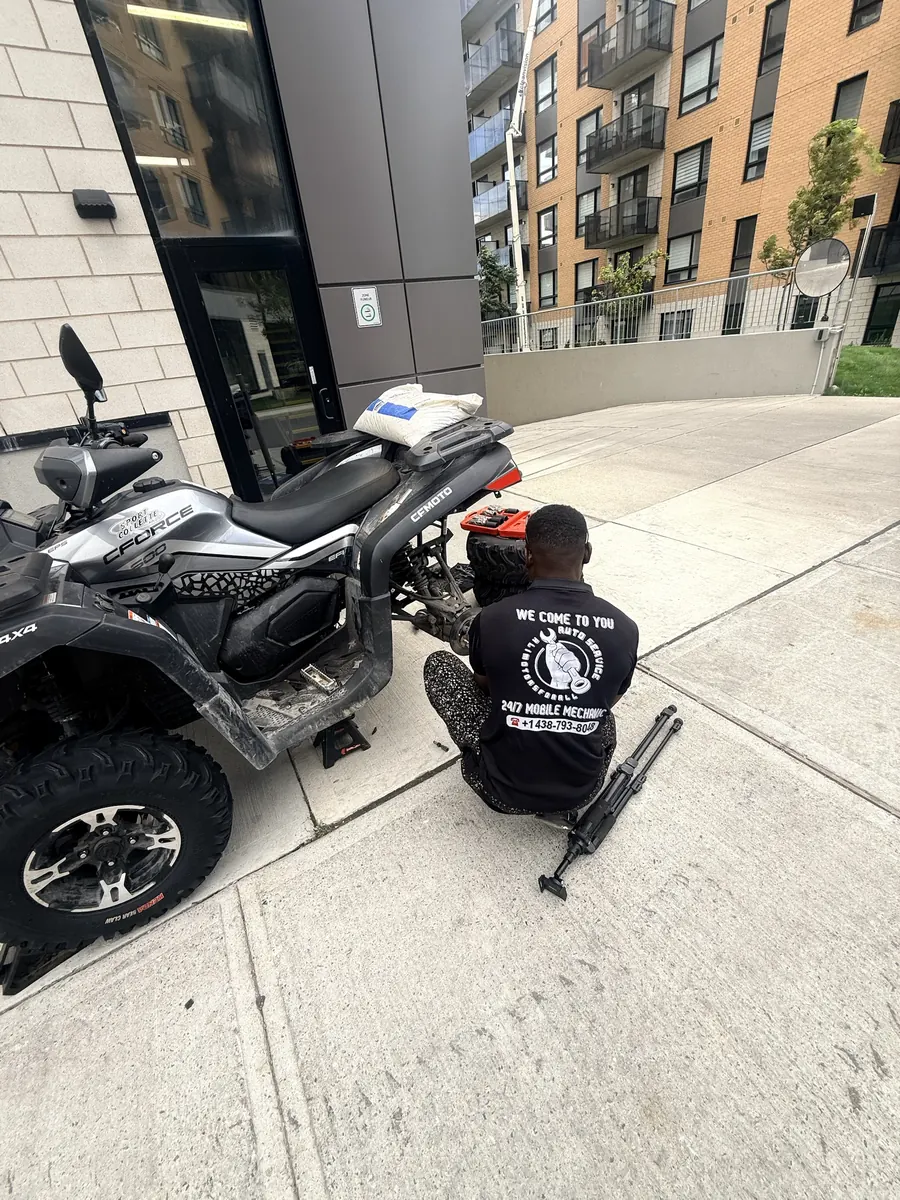 Elie in an Alimotorsforall shirt working at the rear wheel of a CFMoto CForce ATV beside an apartment building