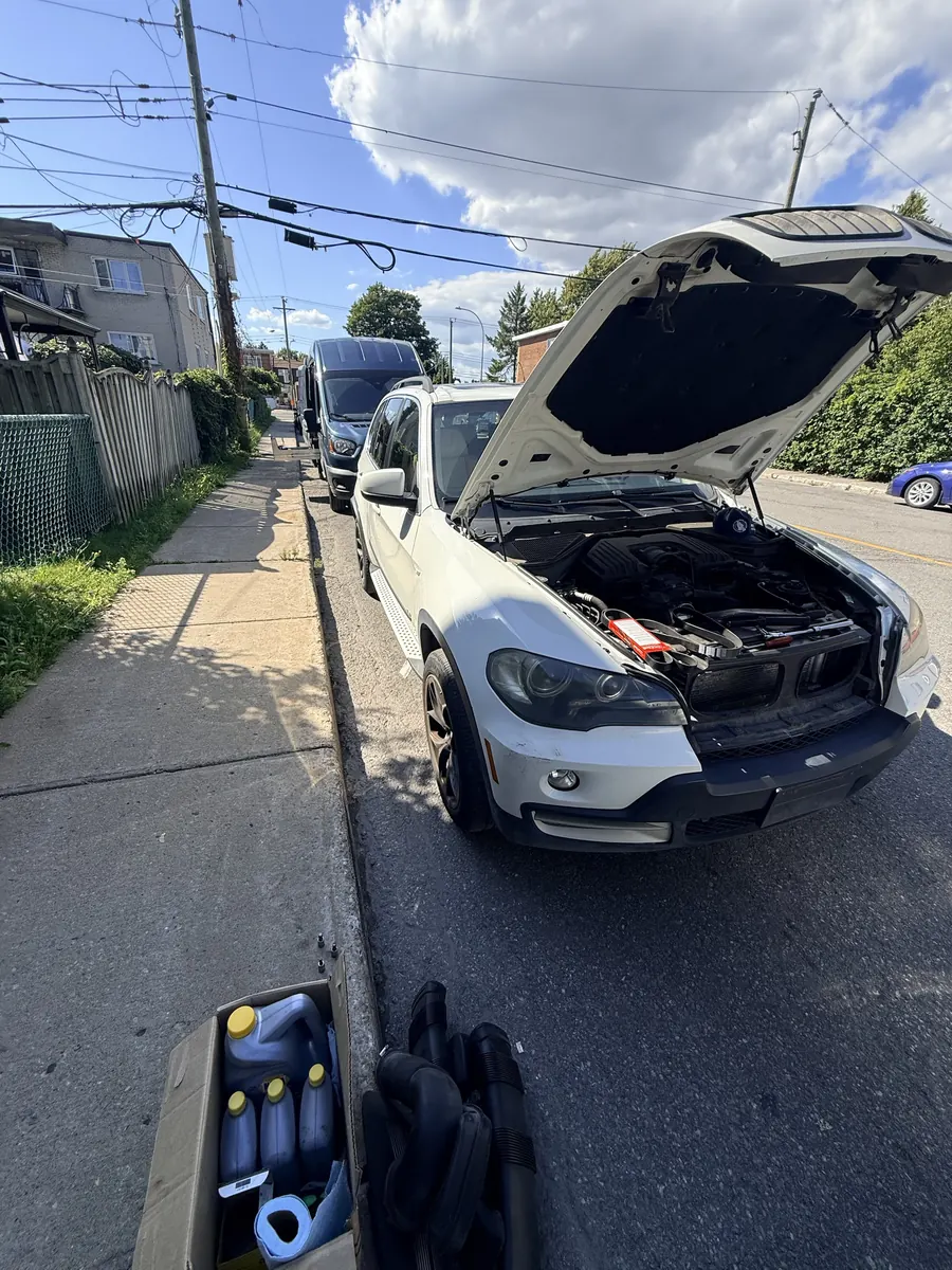 White BMW X5 with the hood open on a residential street, oil and parts in a box on the sidewalk