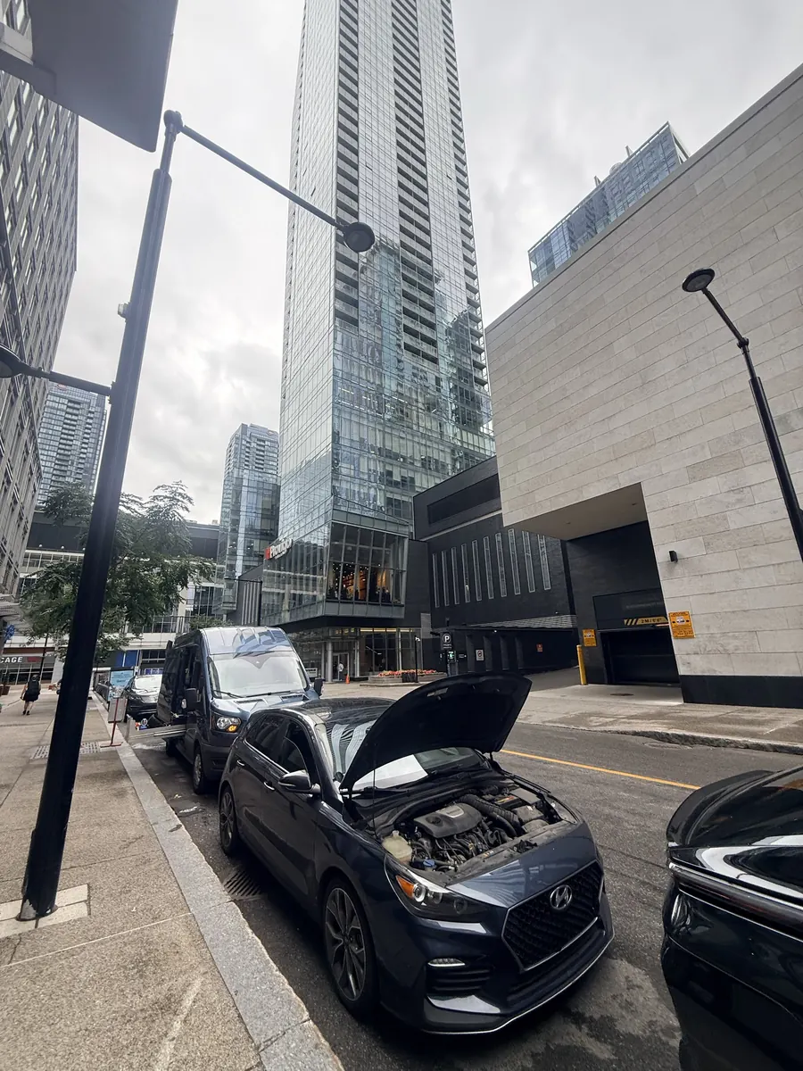 Hyundai hatchback with the hood open at the curb on a downtown Montreal street, blue Ford Transit service van parked behind it