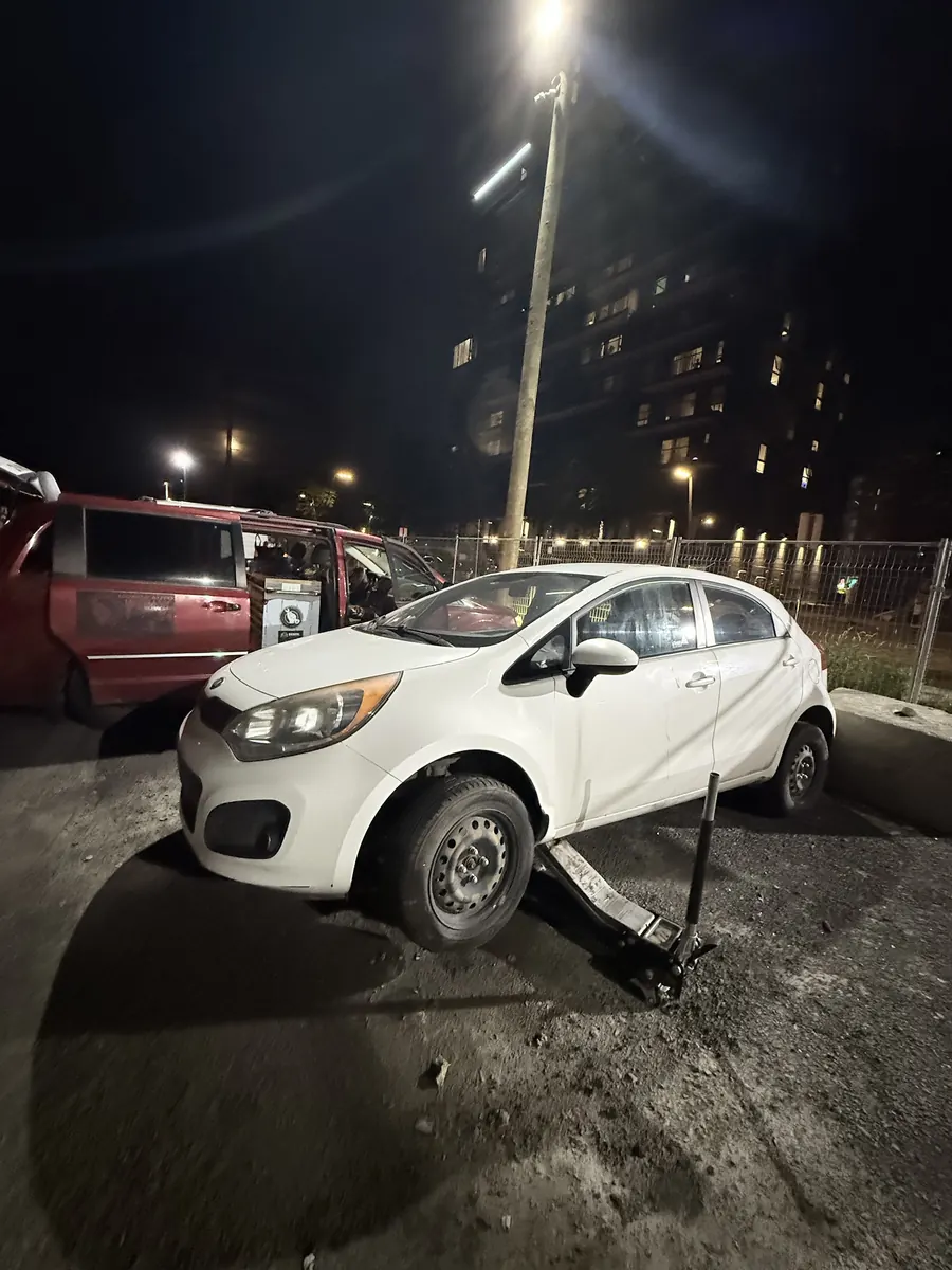White Kia Rio raised on a floor jack in a parking lot at night under a street light, service vehicle beside it