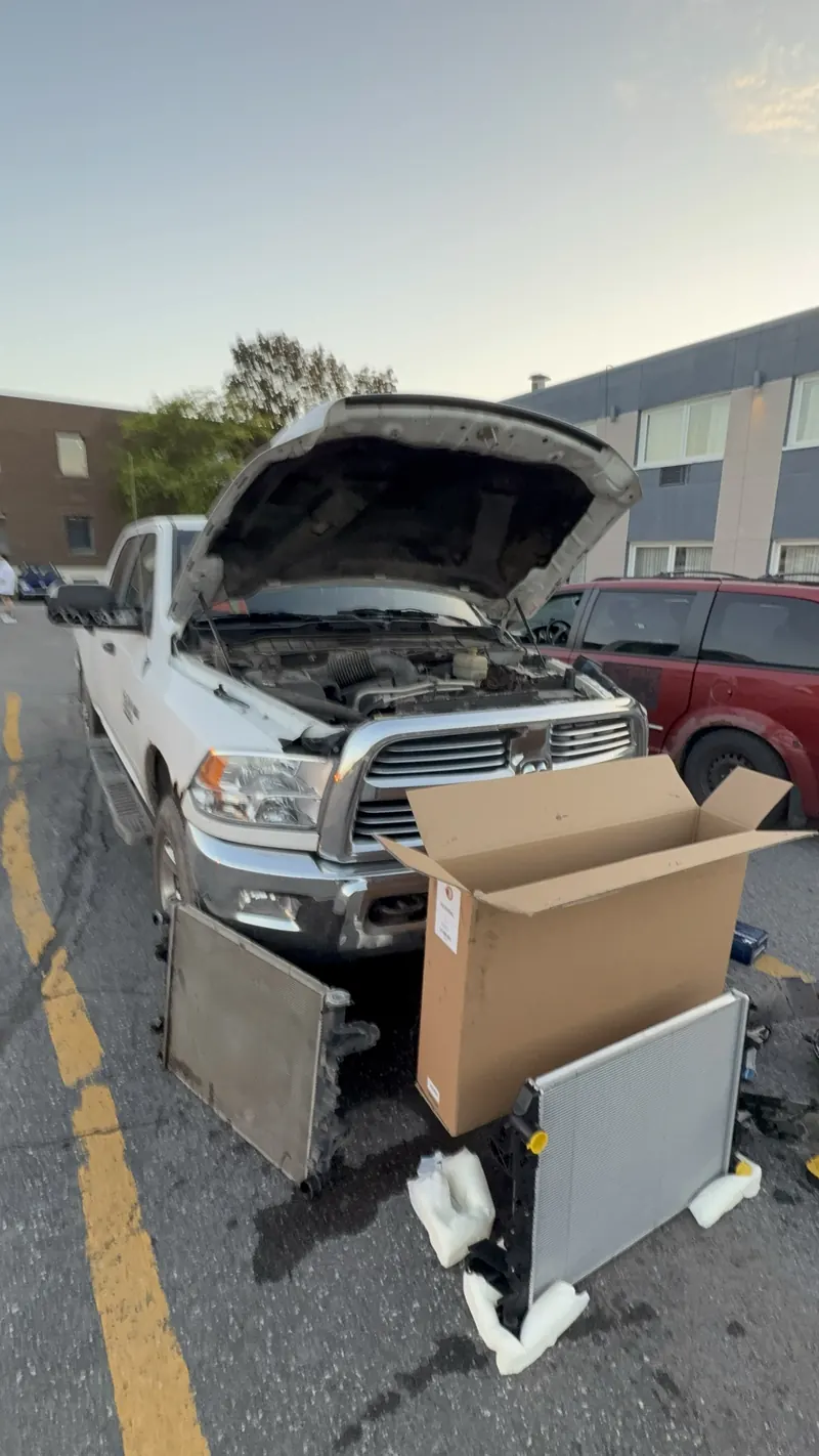 White Ram pickup with the hood open, old radiator removed and new radiator unboxed in a parking lot