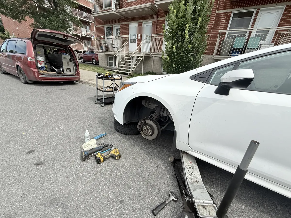 White car on a floor jack with the front wheel off on a residential street, service minivan behind