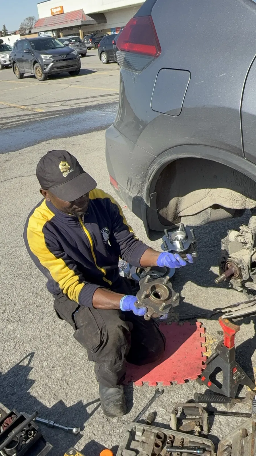 Elie holding the worn wheel hub and the new hub bearing next to a grey Nissan Rogue